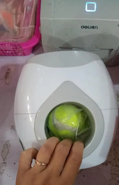Smart pet feeder reward machine with two tennis balls and treat dispenser in green and white colour, shown on a wooden floor