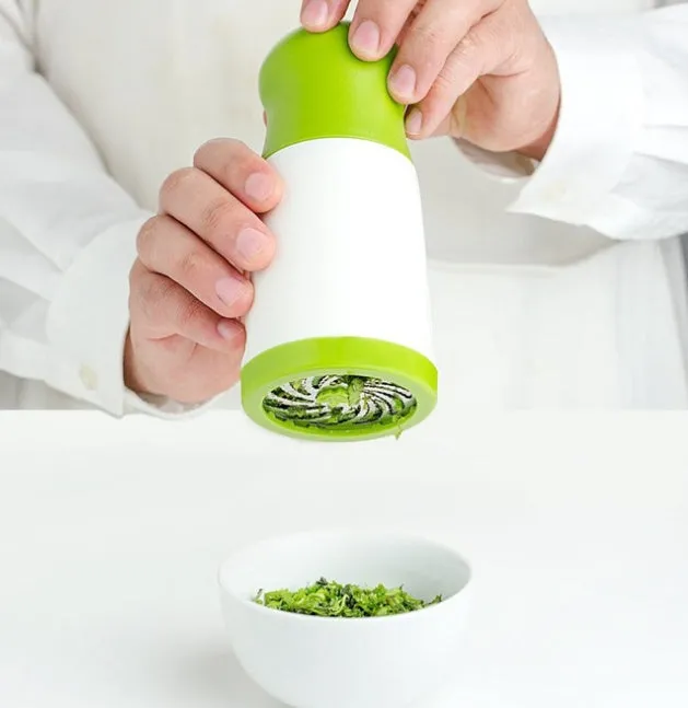 White and grey manual herb grinder with stainless steel blades and removable top on a kitchen counter.