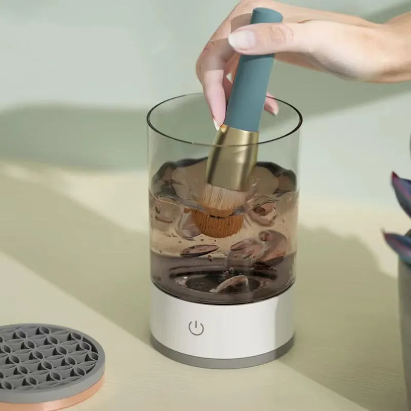 Grey and white electric makeup brush cleaner with USB charging cable on a wooden surface