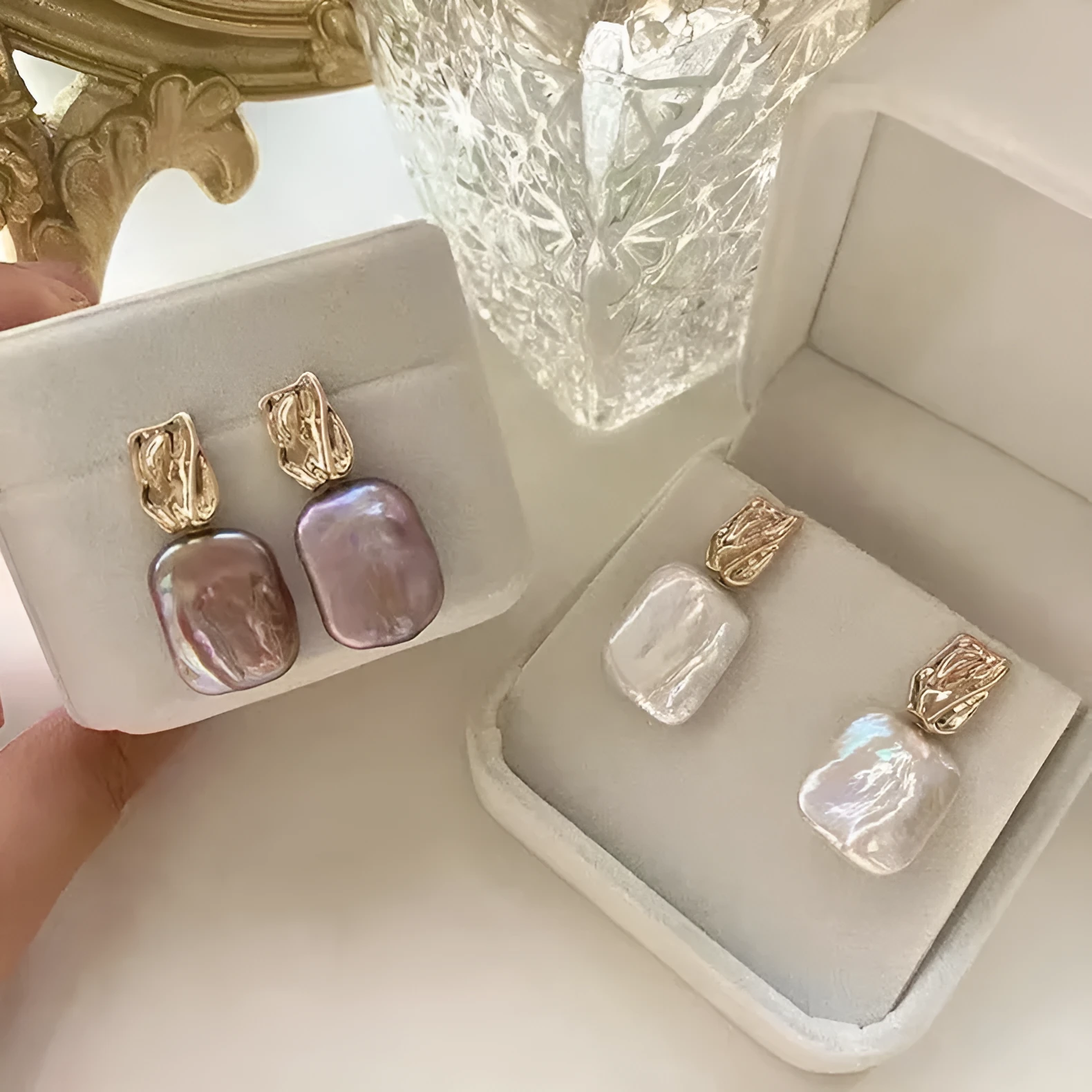 A pair of square stud earrings with a natural freshwater pearl set in a geometric alloy frame, shown in purple.