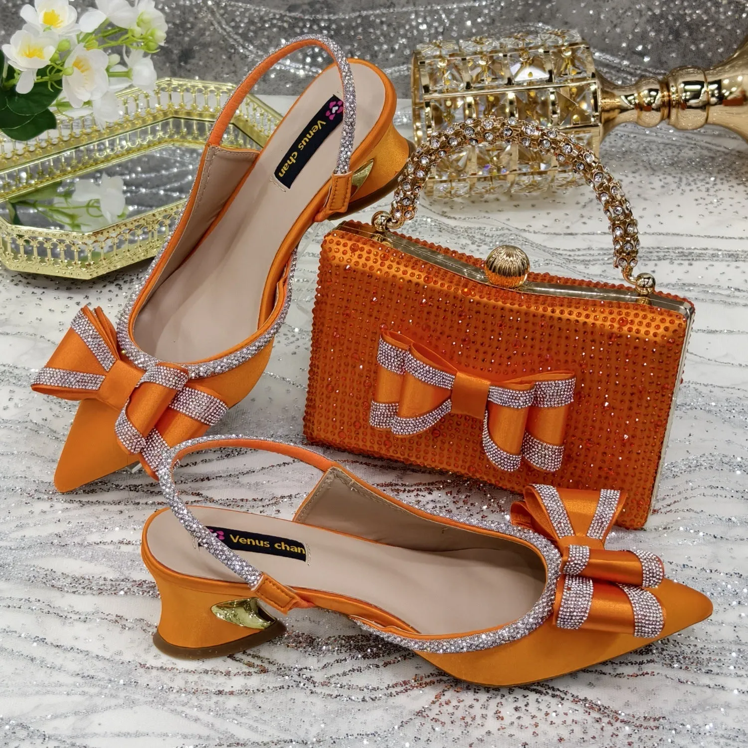 Women's black pointed sandals with a square rhinestone buckle and mid heel, next to a matching clutch bag.