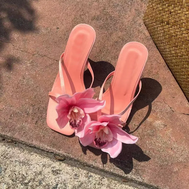 Pink women's sandals with a satin upper, artificial white Phalaenopsis flower, square toe, and a mid heel.