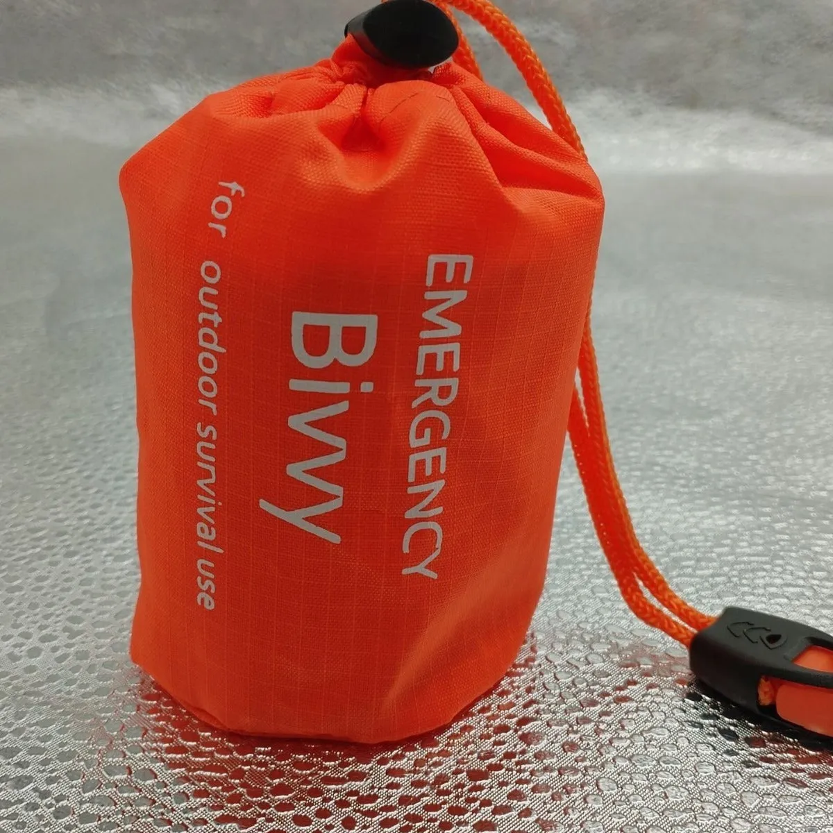 Orange aluminium foil emergency sleeping bag with drawstring bag and whistle on ground
