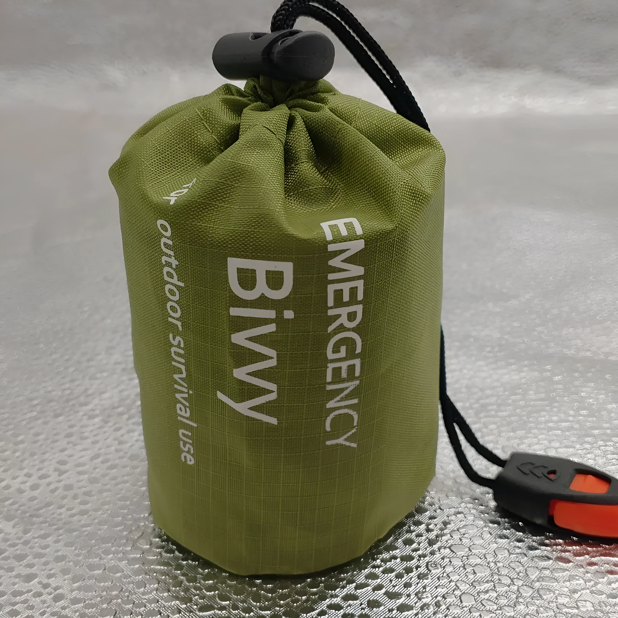 Orange aluminium foil emergency sleeping bag with drawstring bag and whistle on ground