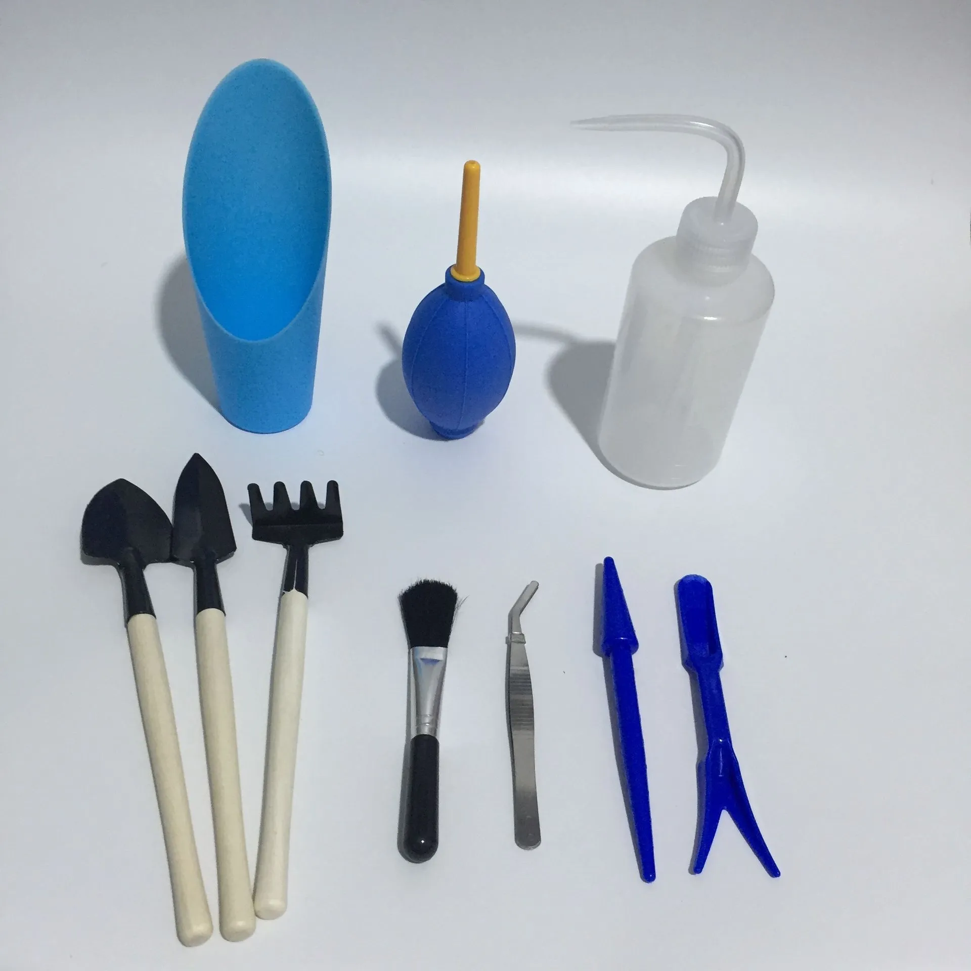 Succulent gardening tool set showing a small watering can, bucket, mini shovel, rake, tweezers, and scissors on a table.