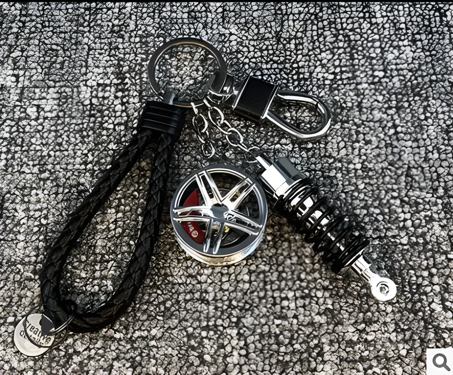 Silver metal wheel and shock absorber keychain with black braided rope and waist buckle on a white surface