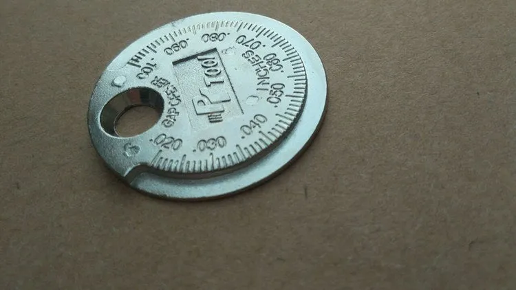 Round zinc alloy spark plug gauge with dual scales on a white background