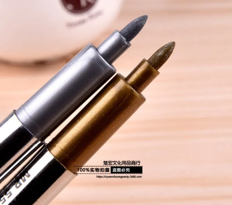 Gold and silver metallic marker pens on a glass surface showing fine tip ink marks